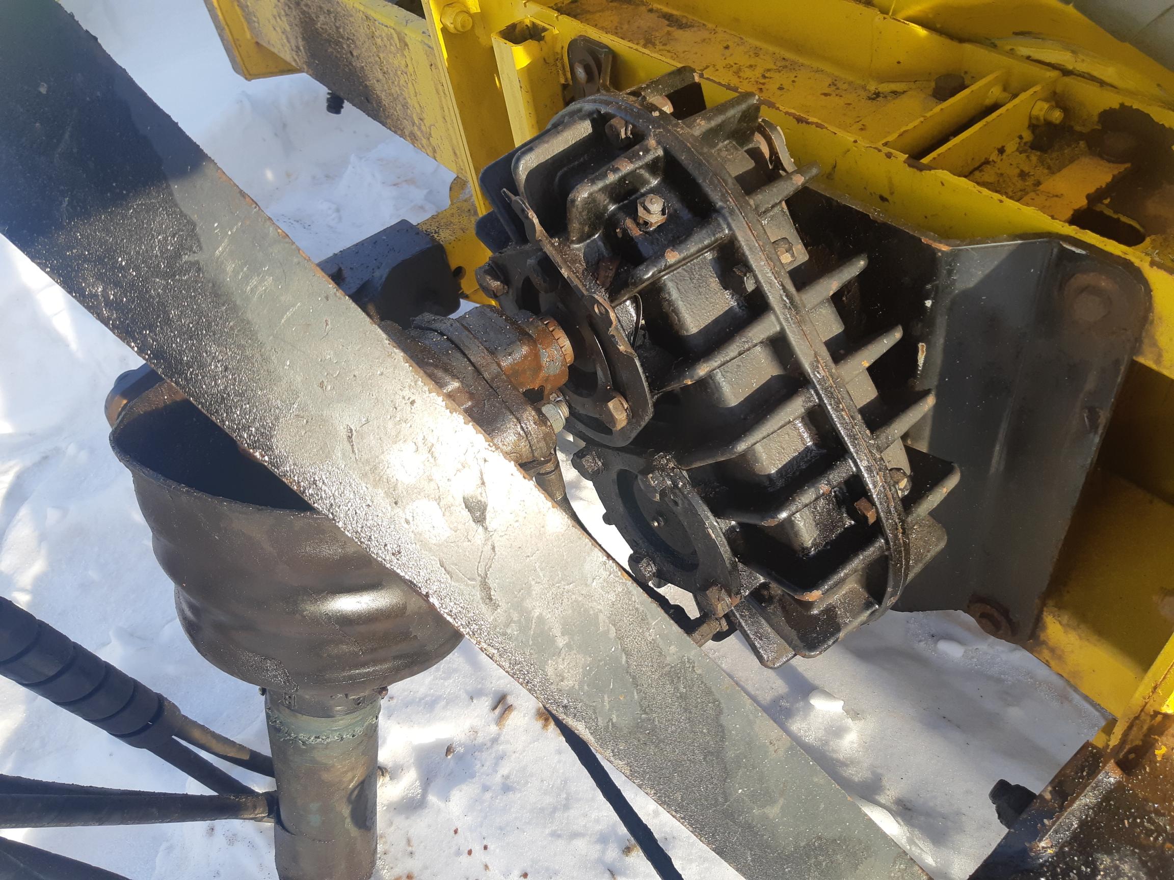 Close-up of industrial machinery with prominent black components, showing a metal bracket connected to a yellow structure. The machine appears to be outdoors, as snow can be seen on the ground and various parts, indicating a cold environment.