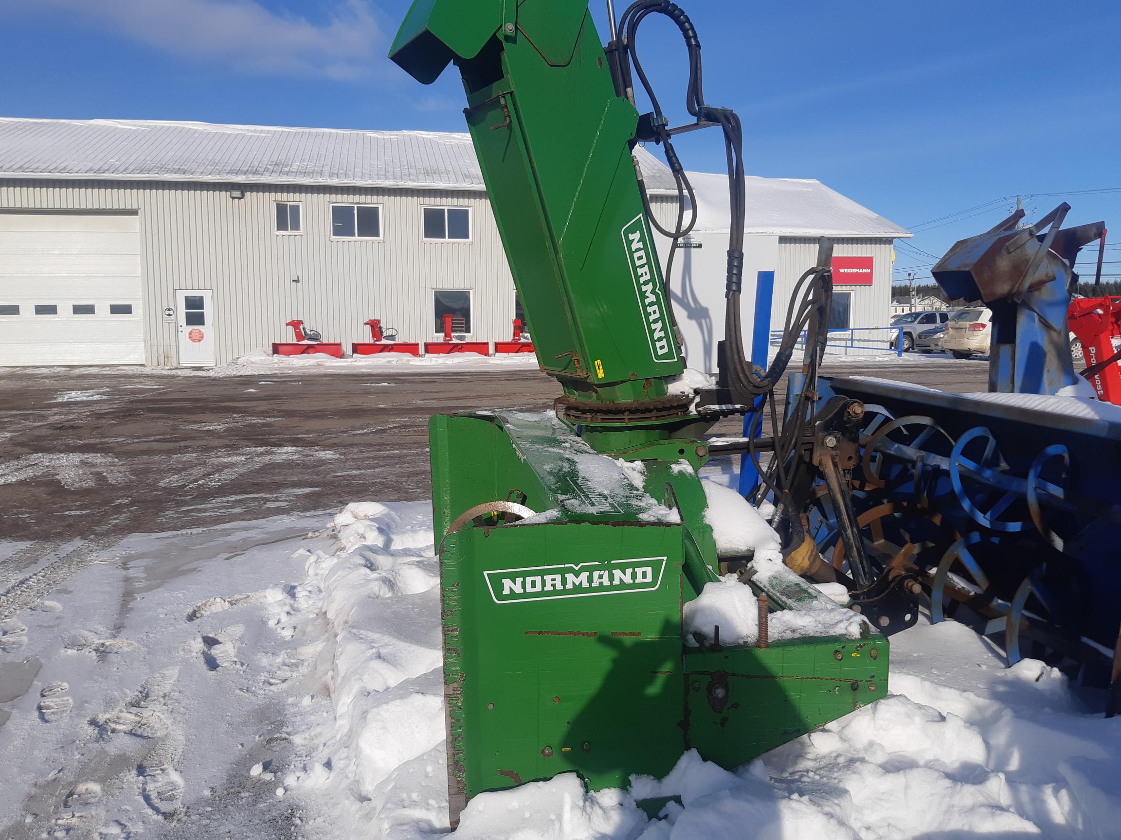 A green Normand snow blower attachment sits on a snowy ground outside a large, white industrial building with several windows and a garage door. Several other pieces of red machinery can be seen in the background.