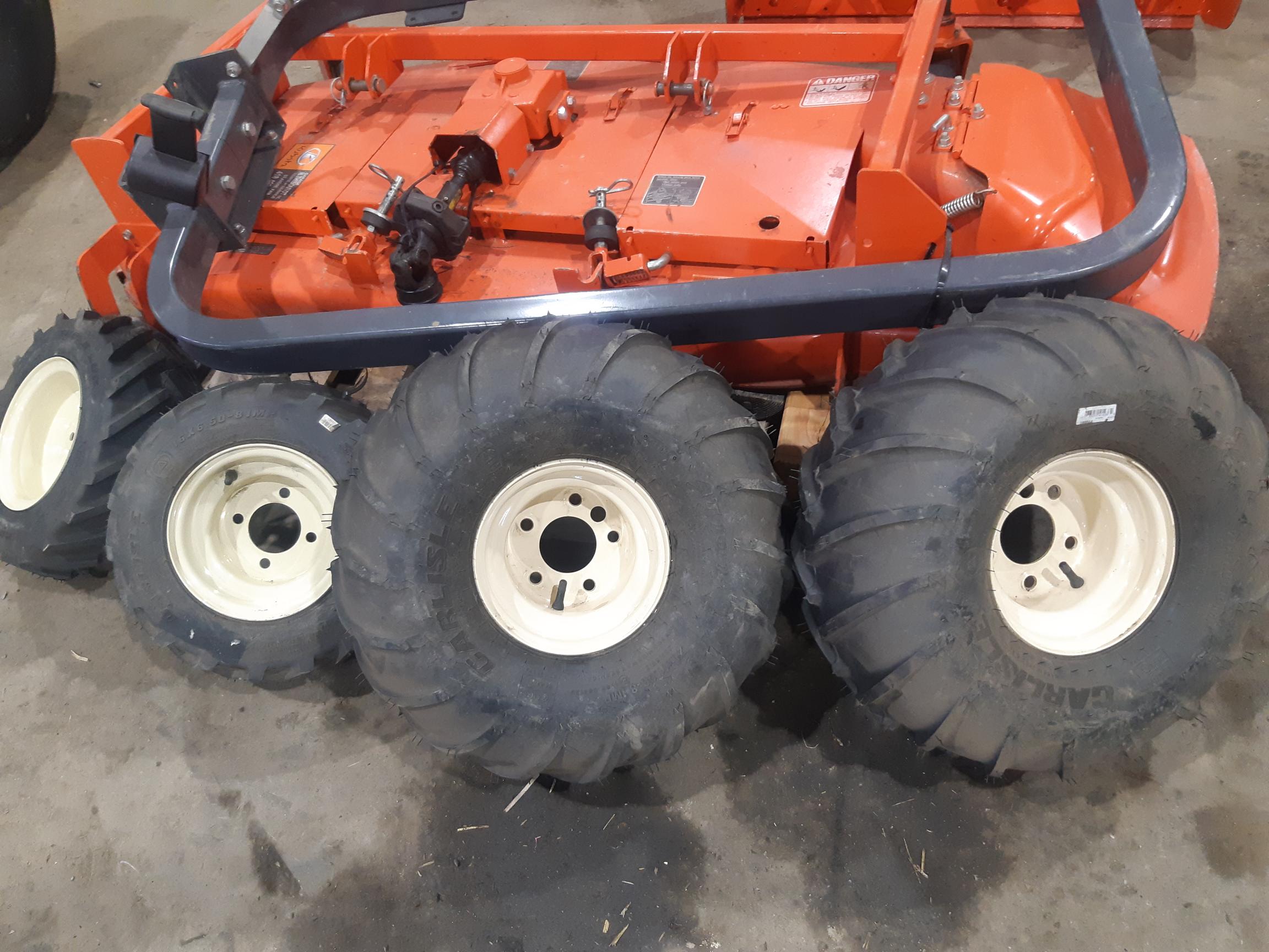 A close-up image showing four large black tires with beige rims lined up on the ground. Behind them, a bright orange agricultural machine is partially visible, placed inside a well-lit garage or workshop with a concrete floor.