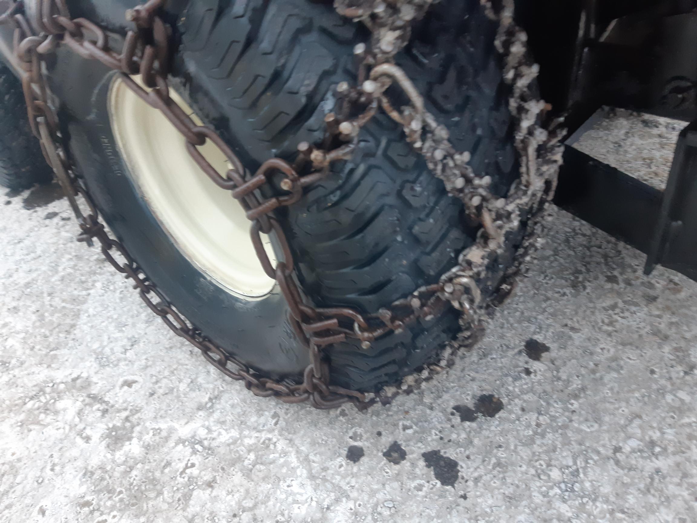 A close-up of a vehicle's tire with snow chains wrapped around it. The tire is on a rough, snowy ground, and the chains are securely fastened to provide traction.