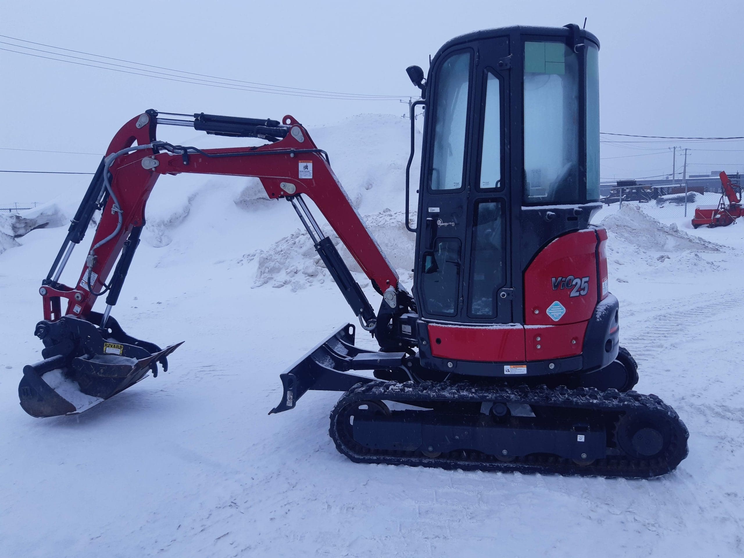 A red and black compact excavator with a fully enclosed cab is parked on a snowy ground. The excavator has a digging bucket attached to its arm, which appears slightly raised. Snowy mounds and other construction equipment are visible in the background.