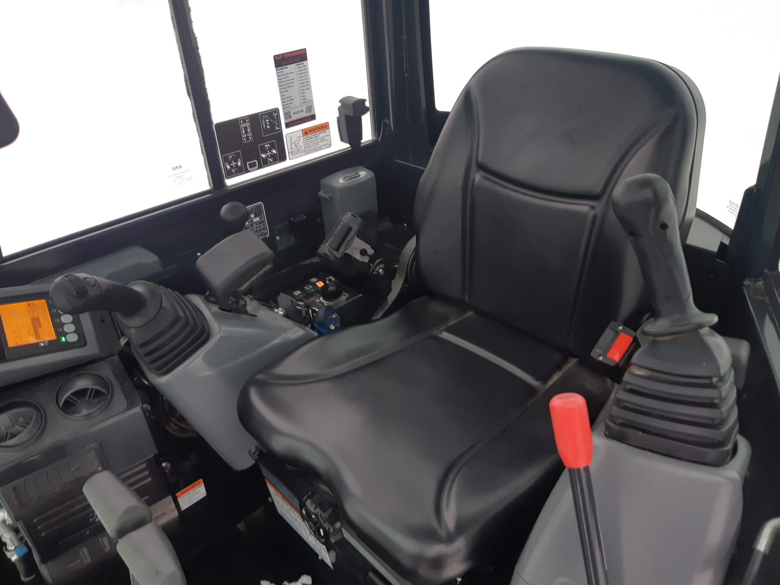 Interior of a construction vehicle cab with a large black cushioned seat. The dashboard includes various controls, levers, and buttons. The left side shows a small digital display, while the right side has two large joysticks with a red lever next to the right joystick.