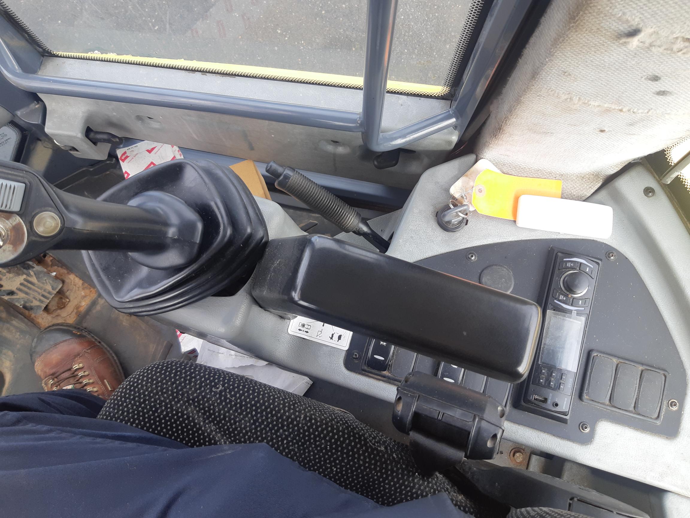 An interior close-up of a vehicle's control console, showing a gear lever, various switches, buttons, and a section of the steering column. A person's leg and shoe are visible at the bottom left corner, seated on a fabric-covered seat.