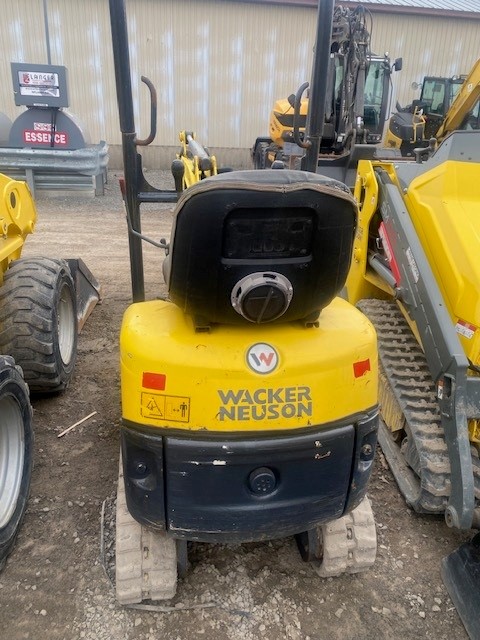 A yellow Wacker Neuson compact excavator with a black driver's seat, viewed from the rear, is parked on a dirt lot. Nearby, there are more construction vehicles, including another yellow equipment piece on the right and a tire loader on the left. An industrial building is in the background.