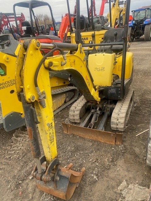 A yellow Wacker Neuson 803 mini excavator is parked in a lot filled with various construction equipment. The excavator has tracks and a front blade with an extended arm featuring a bucket attachment. Other machinery is visible in the background.