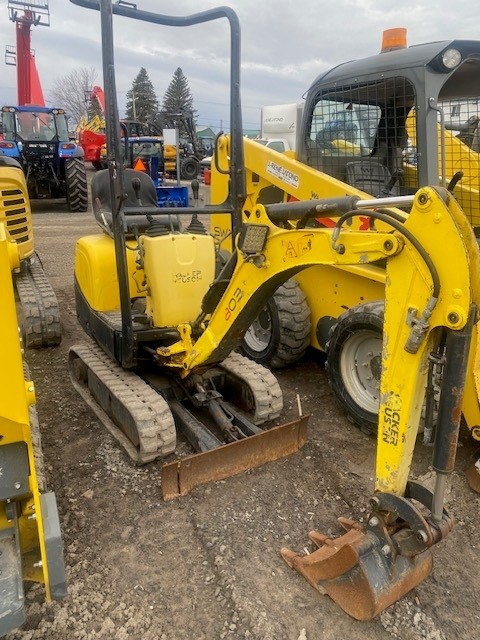 A small yellow mini excavator with a front bucket and arm attachment is parked on a gravel surface among other construction vehicles. The excavator has 