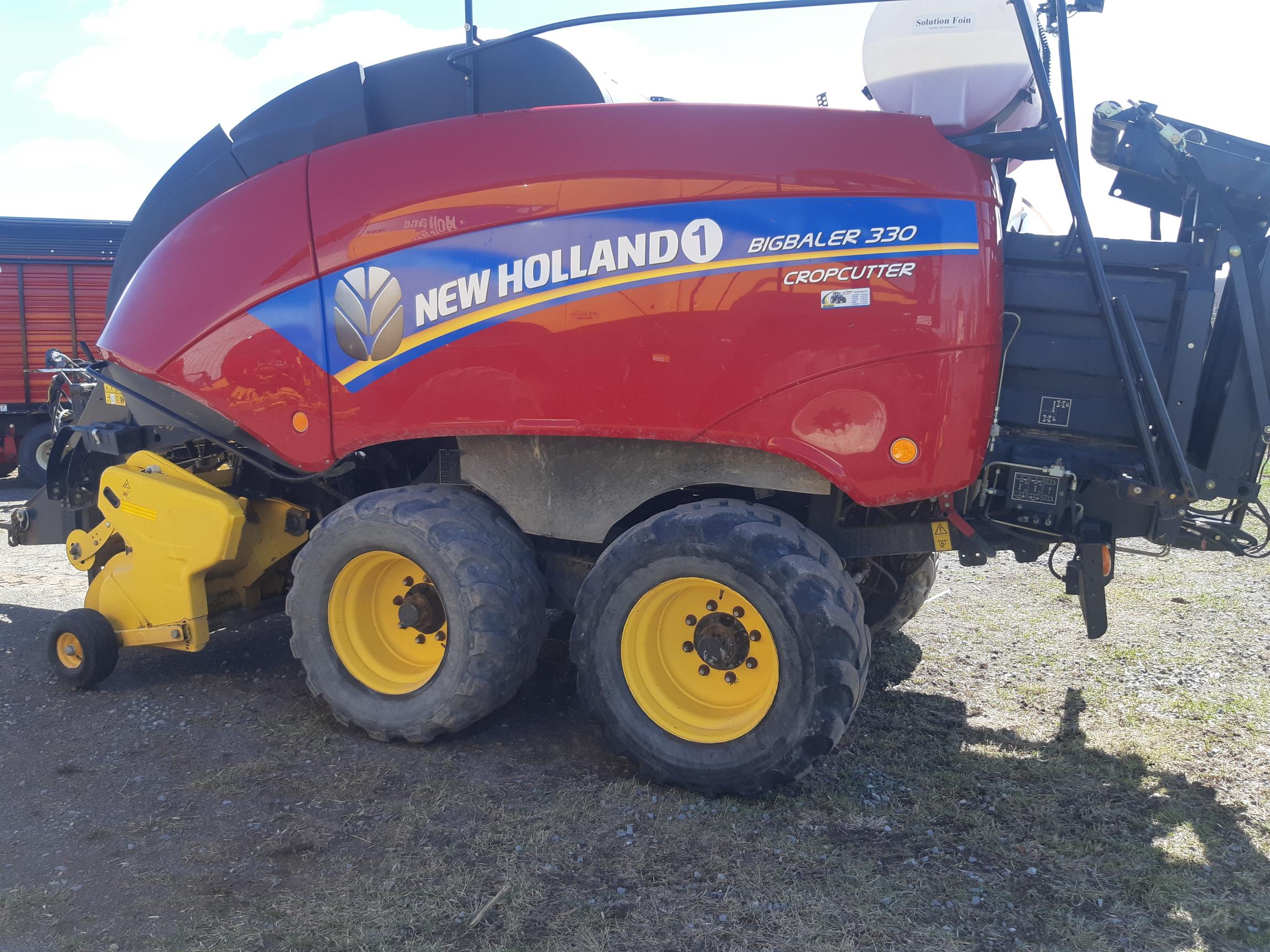 A New Holland BigBaler 330 CropCutter baler with a red and yellow body and large black tires is parked on grassy ground on a sunny day. The baler has visible branding and logos on its side.