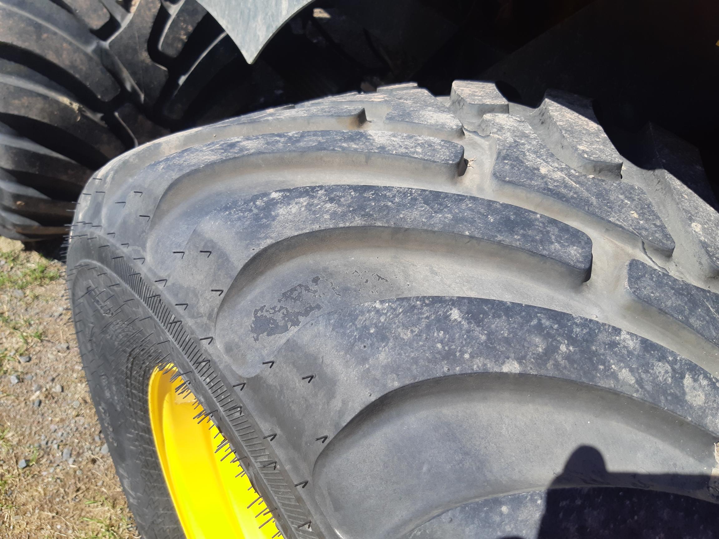 Close-up view of a large, rugged off-road tire with deep, pronounced treads. The tire is mounted on a wheel with a yellow rim. Dirt and some wear are visible on the tire surface, and a partial shadow appears in the bottom right corner.