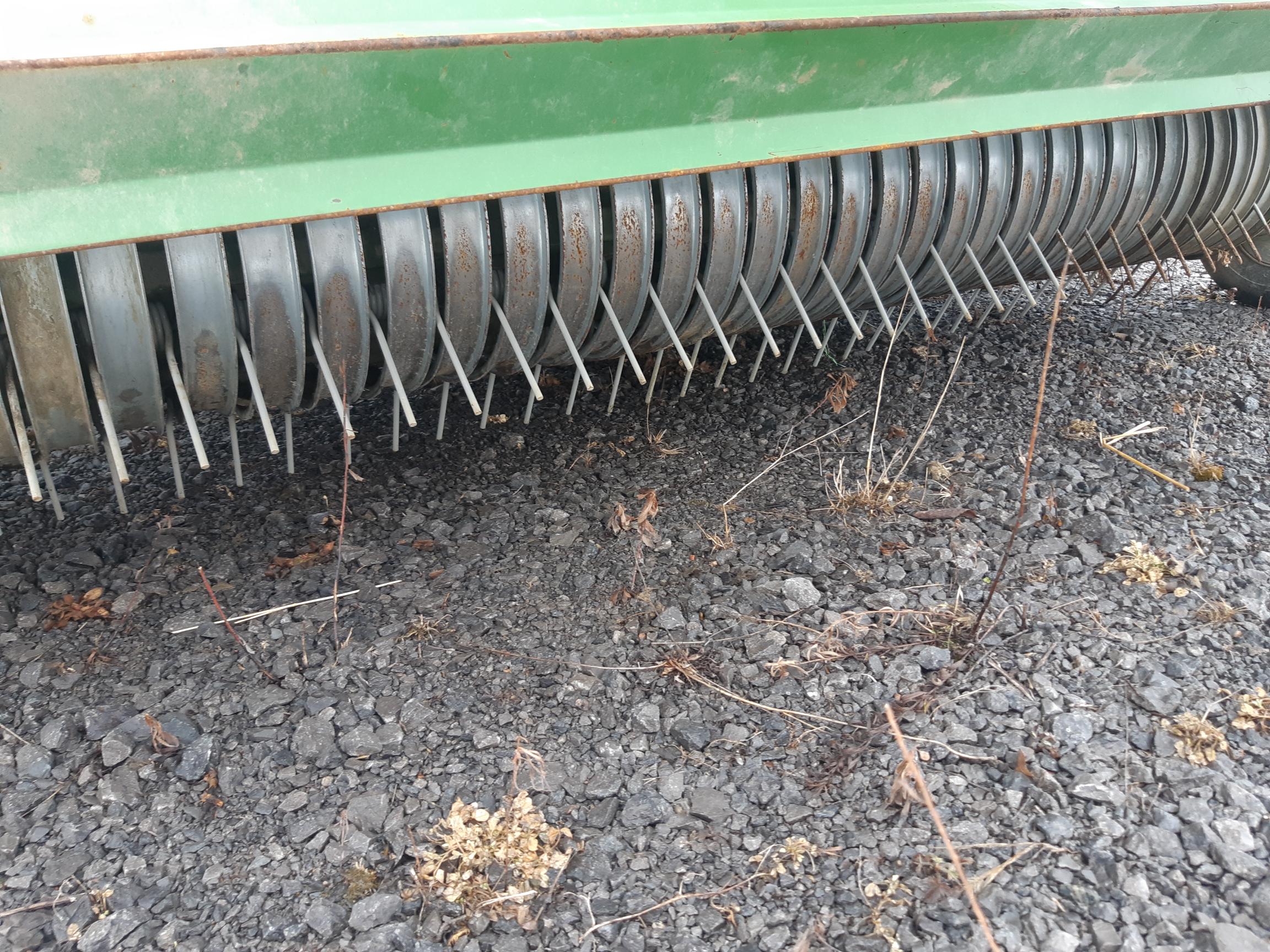 Close-up view of the underside of a green agricultural machine, showing multiple metal spinning tines used for tilling or raking the ground. The tines are over a gravelly and weed-strewn surface.