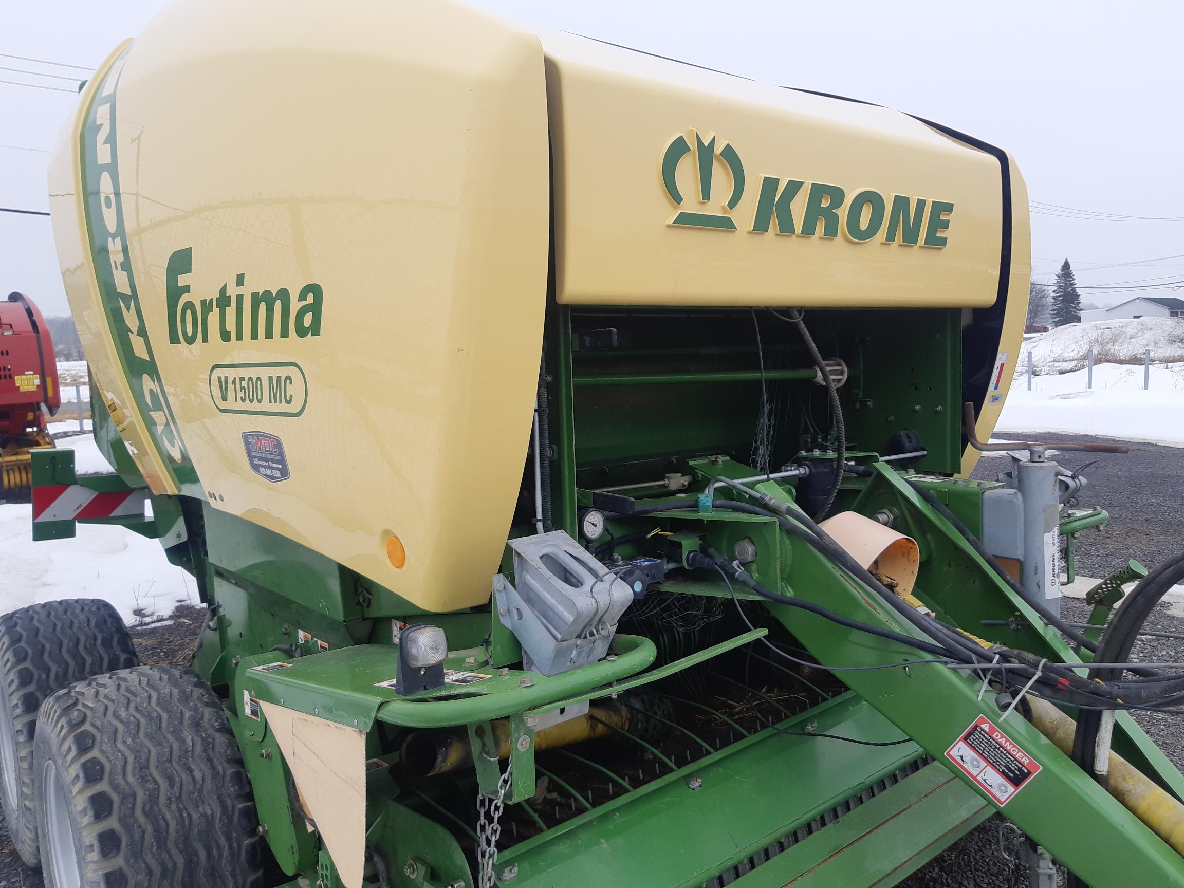 Close-up of a Krone Fortima V1500 MC round baler machine, predominantly yellow with green accents, equipped with large, textured tires. It is situated outside on a cold, overcast day, with snow visible in the background.