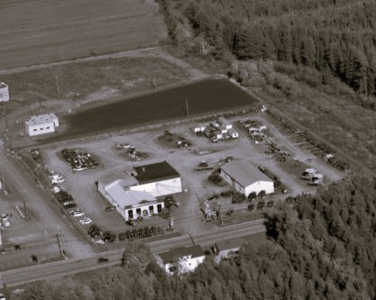 Vue aérienne d'une propriété rurale près de Rivière-du-Loup, comprenant plusieurs grands bâtiments, des voitures stationnées et des champs ouverts entourés d'arbres denses. Une machine New Holland est visible sur le site. L'image est en noir et blanc.