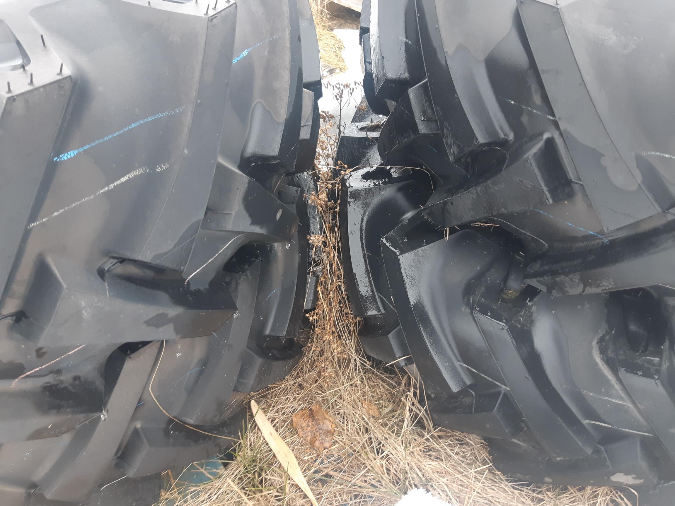 Close-up view of two large black tractor tires with deep treads, placed side by side. The space between the tires reveals dry grass and twigs on the ground. The surface of the tires shows signs of outdoor exposure, with some dirt and scratches visible.