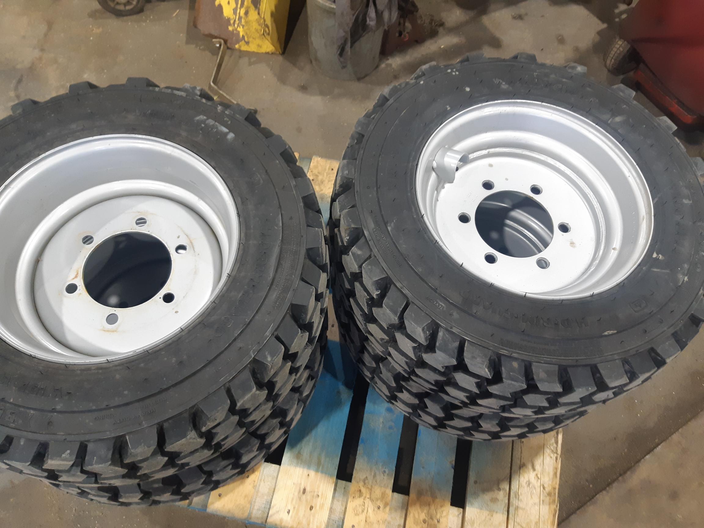 Two heavy-duty tires with white rims are positioned upright on a blue wooden pallet. The tires have deep treads, suggesting they are designed for off-road or industrial vehicles. The background shows part of a workshop with various equipment and tools.