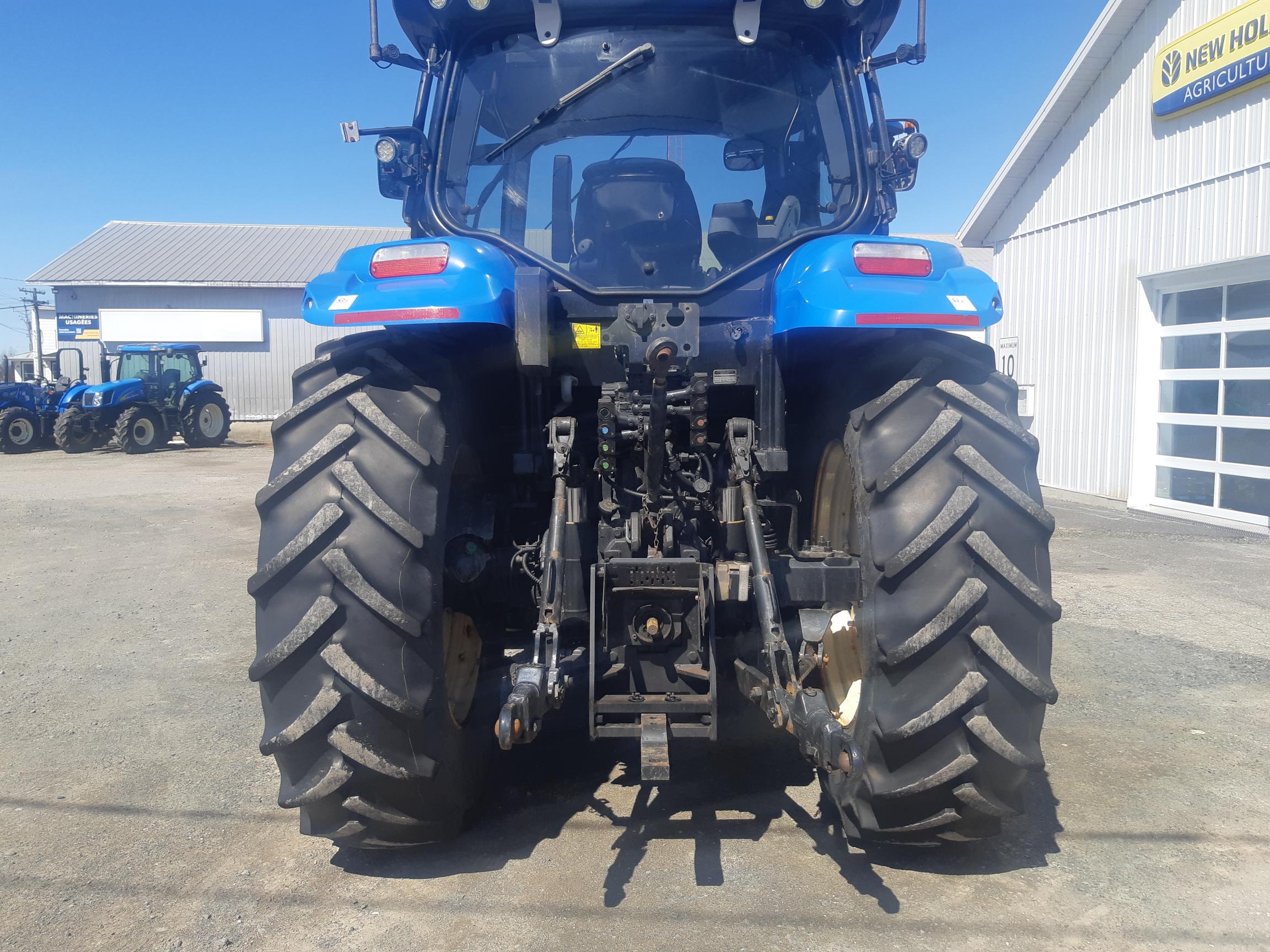 Rear view of a blue New Holland tractor with large, thick tires, parked in a gravel lot outside a white building labeled 