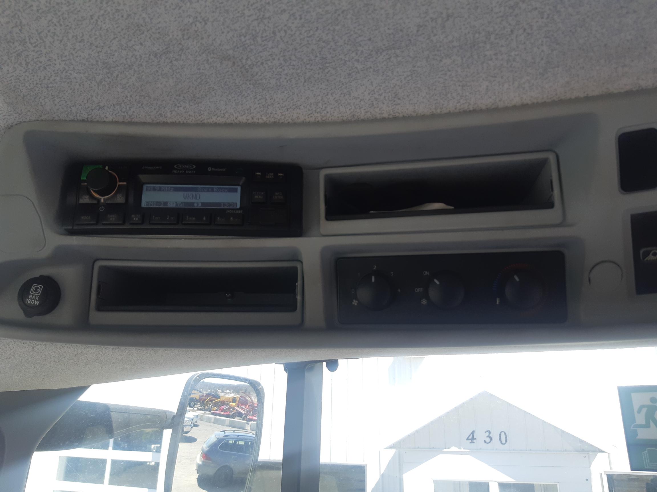 The image shows the interior roof panel of a vehicle, featuring a built-in stereo system, air vents, and three control dials. A window is visible in the lower part of the image, revealing a building labeled 