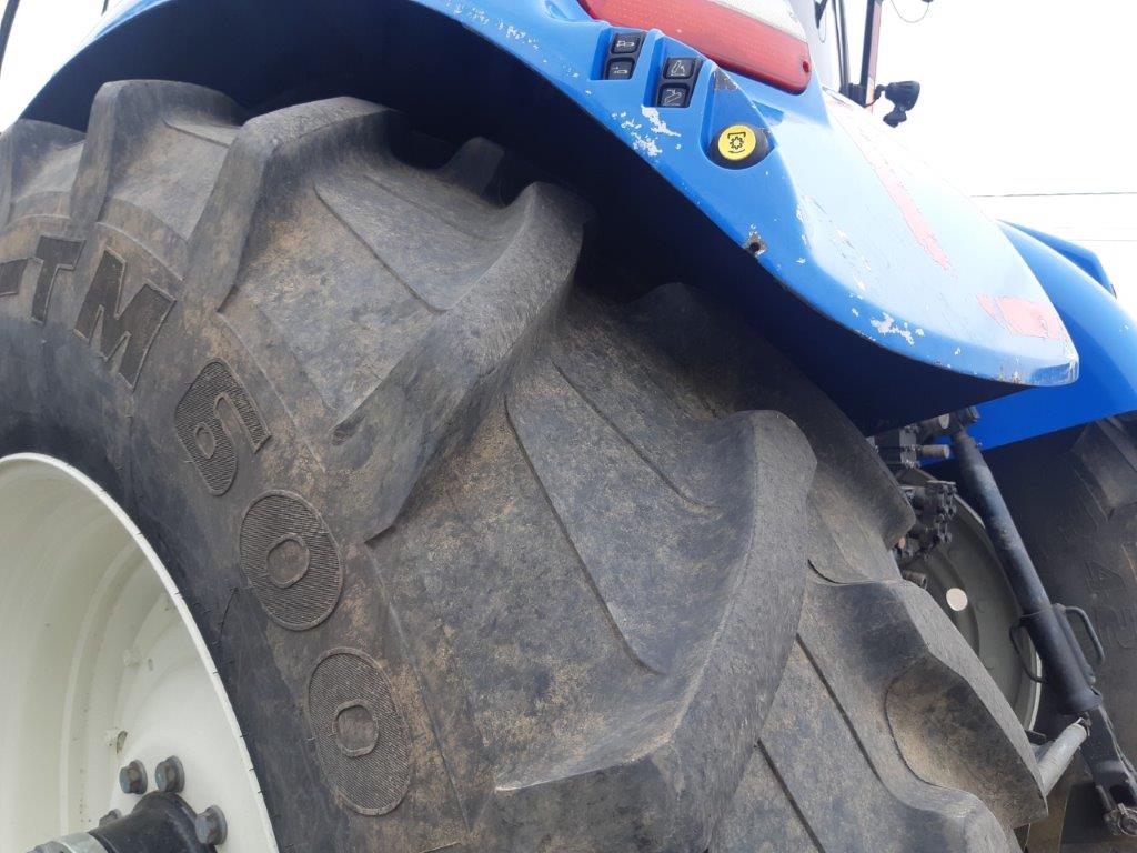 A close-up view of a large, rugged tractor tire showing the tread pattern and part of the blue chassis. The tire has markings 