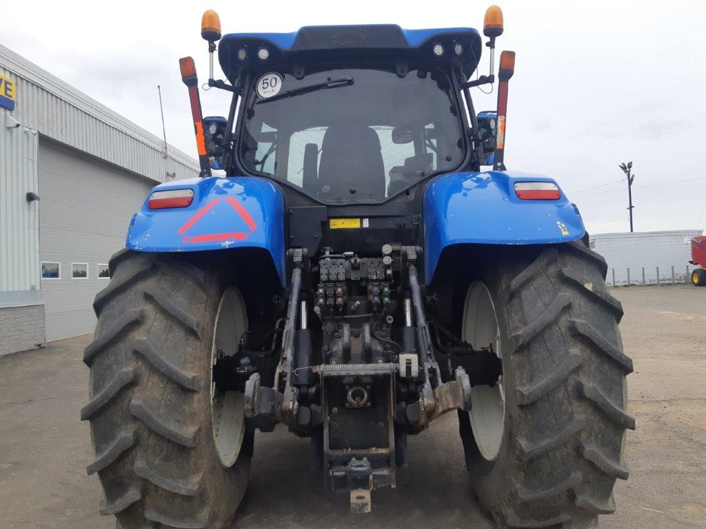 Rear view of a blue tractor parked outdoors. The tractor features large rear tires with visible treads and a triangle safety sign on the back. The background includes a building with a garage door and other farm equipment.