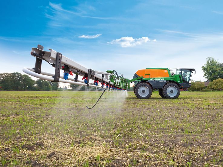 Un grand pulvérisateur agricole automoteur dans un champ, pulvérisant un brouillard de liquide sur le sol. Le pulvérisateur est vert et orange, avec une longue rampe qui s'étend vers l'extérieur. Le ciel est clair et bleu, et il y a des arbres au loin.