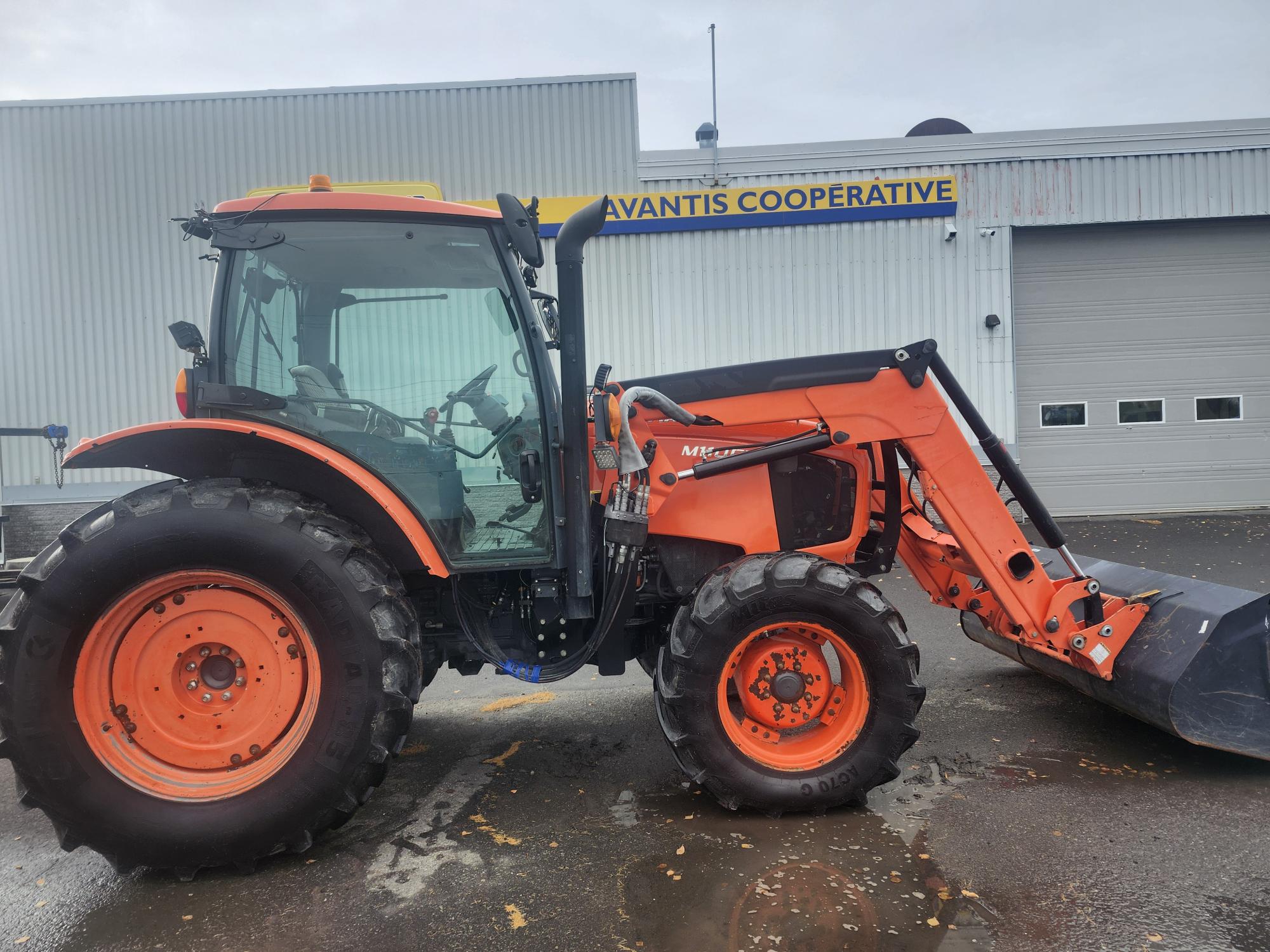 Un tracteur orange équipé d'un chargeur frontal est stationné sur une surface mouillée devant un bâtiment marqué « Avantis Cooperative ». Le tracteur est équipé de gros pneus noirs et de vitres transparentes. Le bâtiment a un extérieur en métal et une porte de garage.