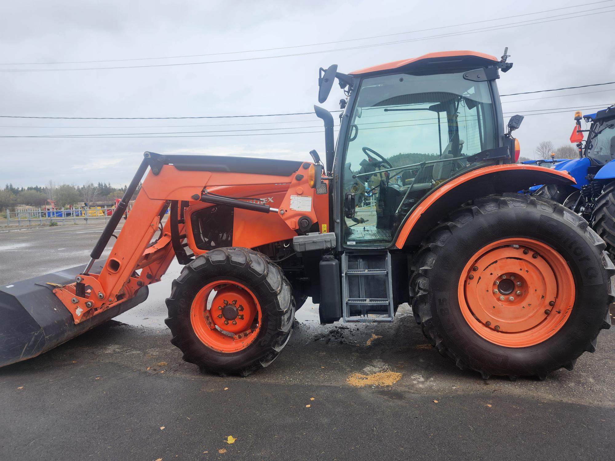 Un tracteur orange équipé d'un chargeur frontal est stationné sur une chaussée mouillée. L'arrière-plan montre un ciel nuageux et quelques lignes électriques. Un autre tracteur bleu est partiellement visible à droite.