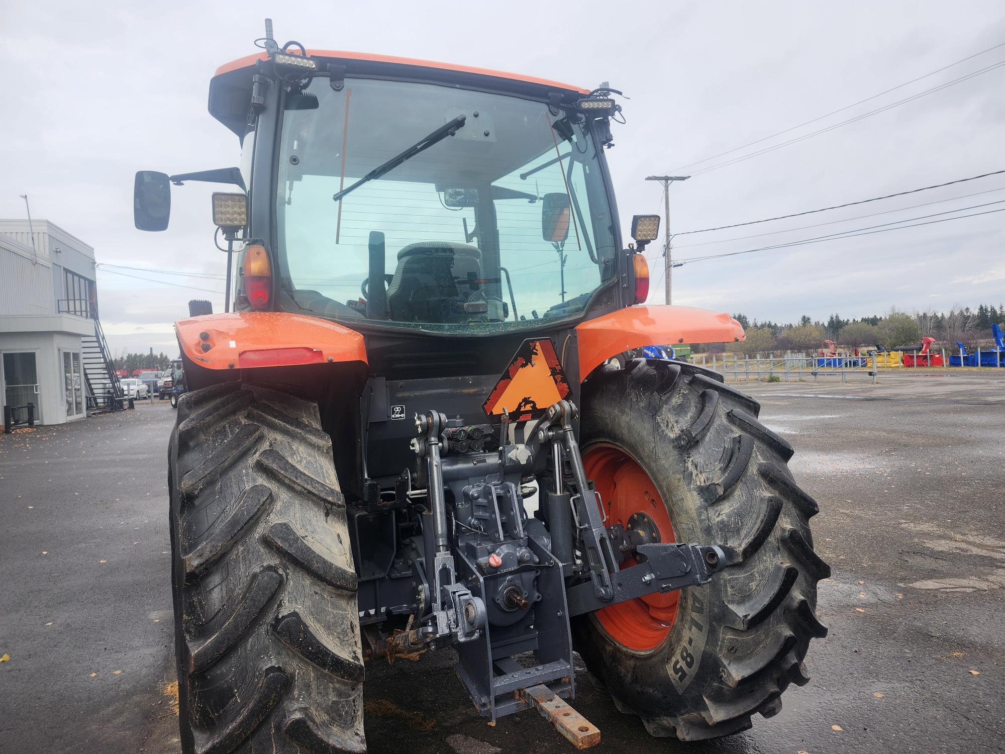 Un gros tracteur orange et noir est stationné sur une surface extérieure mouillée. Il est doté de gros pneus robustes et de divers composants mécaniques visibles à l'arrière. Des bâtiments et des arbres sont visibles à l'arrière-plan sous un ciel nuageux.