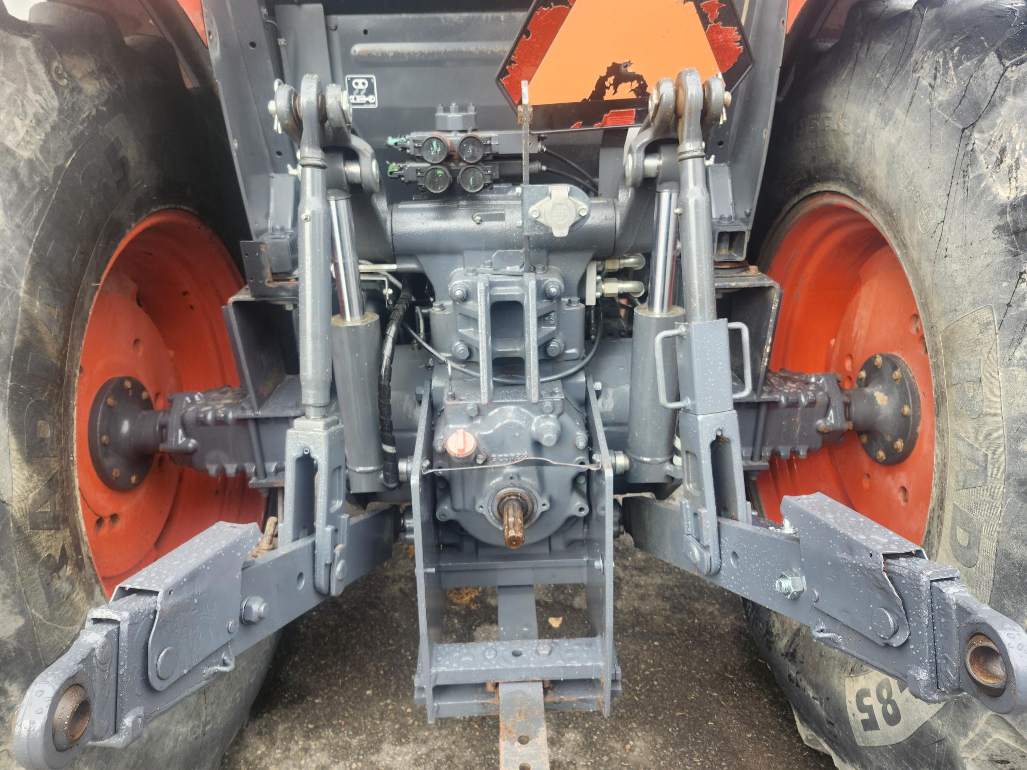 Vue arrière du système d'attelage à trois points d'un tracteur, montrant de gros pneus, des bras hydrauliques et des composants métalliques. La machine semble robuste, posée sur une surface pavée, avec quelques éléments orange et rouillés visibles.