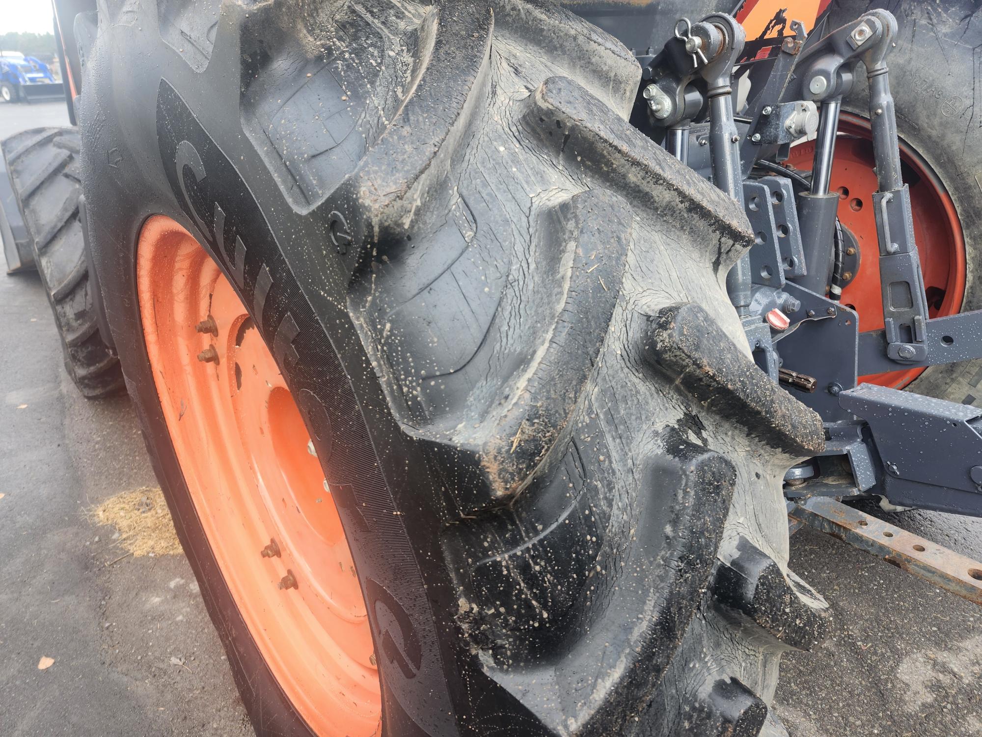 Gros plan d'un pneu de tracteur à bande de roulement épaisse et d'une roue orange sur une chaussée mouillée. Les pièces mécaniques du tracteur sont visibles en arrière-plan. Le pneu présente des traces de saleté et d'usure sur sa surface.