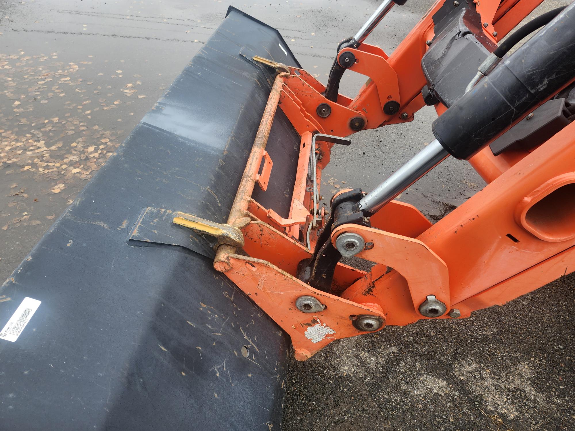 Une rétrocaveuse orange équipée d'un grand chasse-neige noir est positionnée sur une surface en béton humide parsemée de feuilles. Les bras hydrauliques et les composants métalliques sont visibles, indiquant qu'ils sont prêts à déplacer la neige ou les débris.