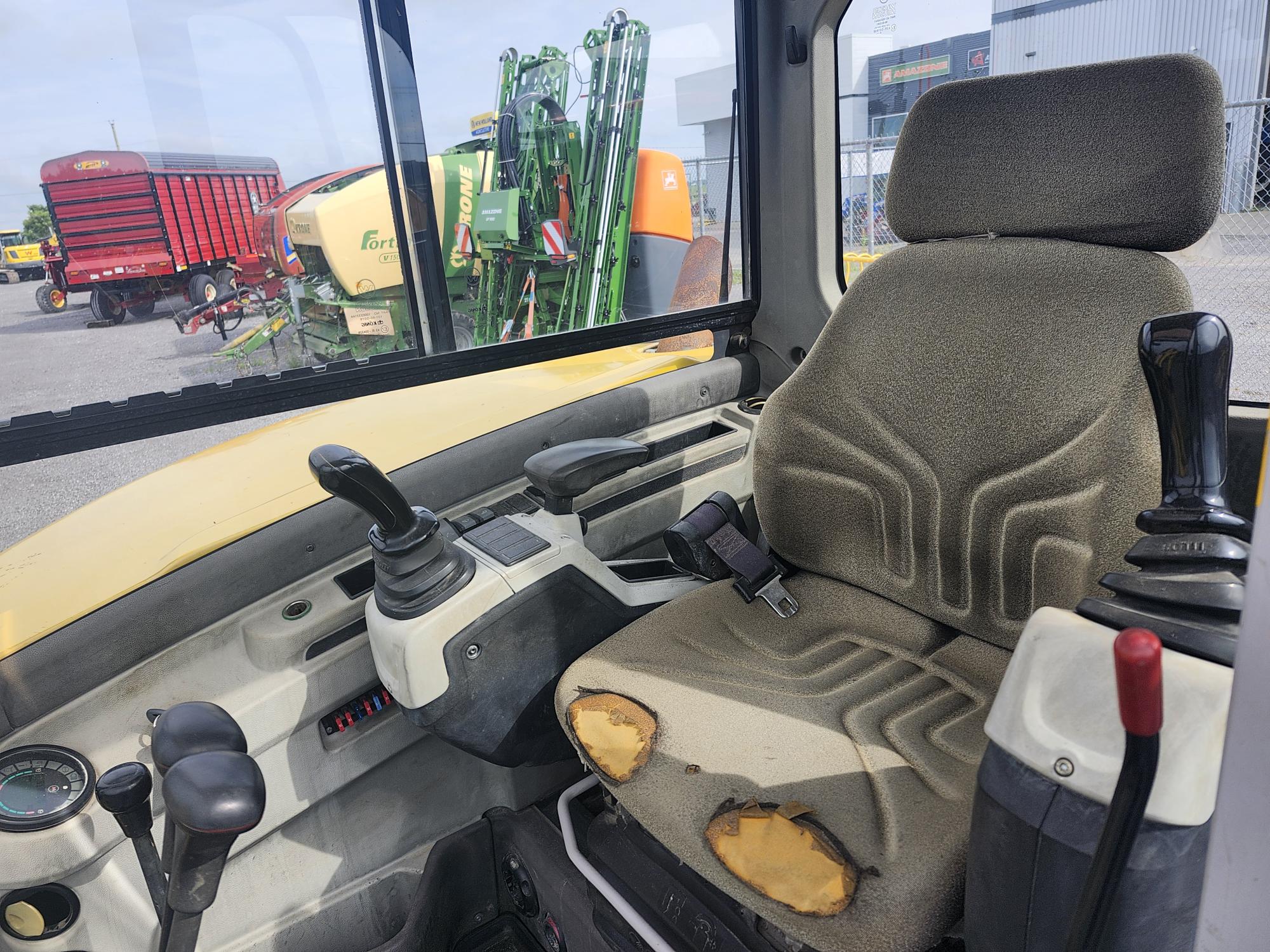 Intérieur de la cabine d'un tracteur montrant un siège conducteur usé avec des déchirures visibles. Les leviers de commande et le volant sont positionnés près du siège. Des machines agricoles et des remorques sont visibles à l'extérieur à travers la fenêtre.