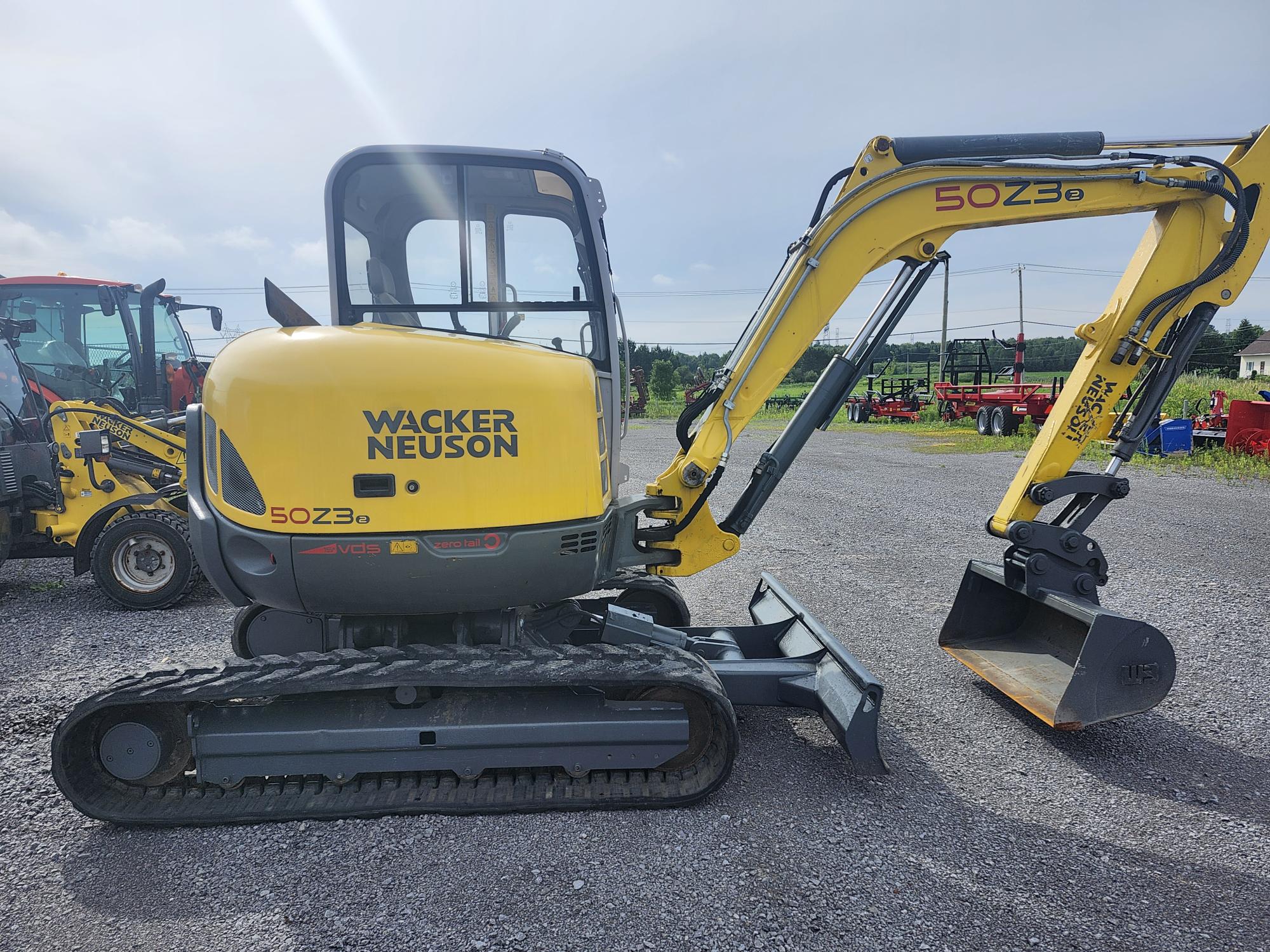 Une pelle compacte jaune Wacker Neuson portant le numéro de modèle 50Z3 est garée sur une surface en gravier. Elle est équipée de chenilles en caoutchouc et d'un godet de creusement. À l'arrière-plan, d'autres véhicules et équipements de construction sous un ciel partiellement nuageux.