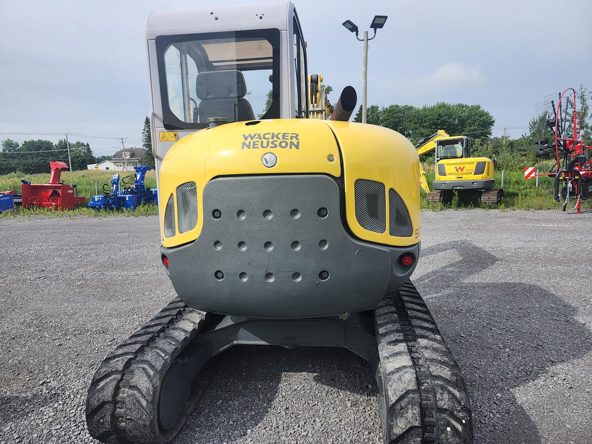 Une excavatrice Wacker Neuson compacte jaune garée sur un terrain en gravier, avec d'autres machines visibles en arrière-plan sur fond de verdure et de ciel clair.