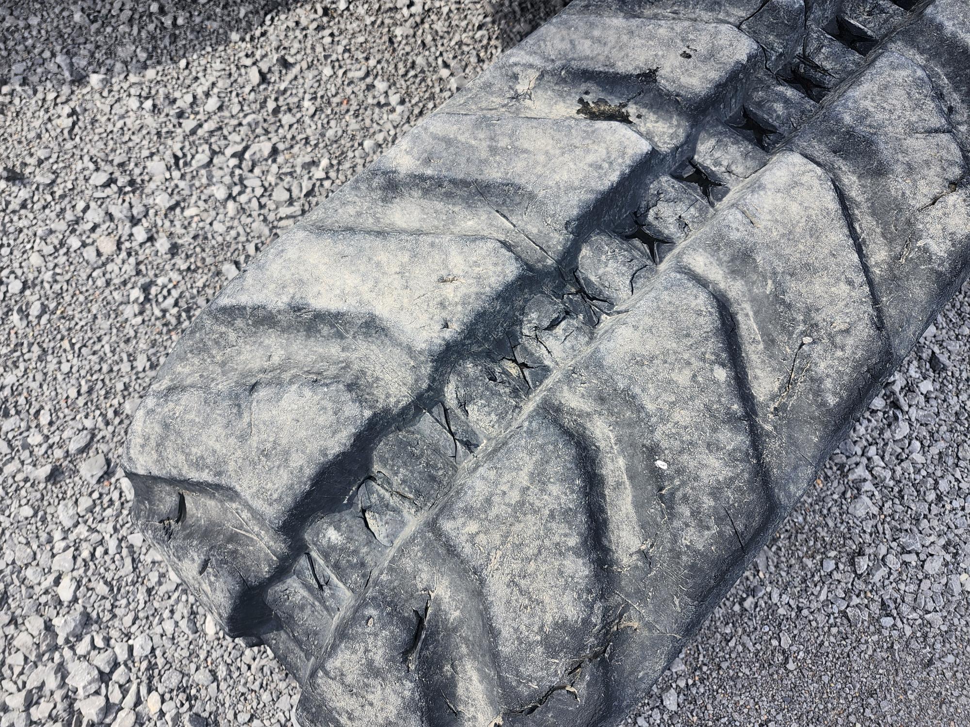 Gros plan d'une bande de roulement usée et fortement fissurée sur un gros pneu de véhicule, posé sur une surface en gravier. La texture du pneu et le sol accidenté sont clairement visibles.