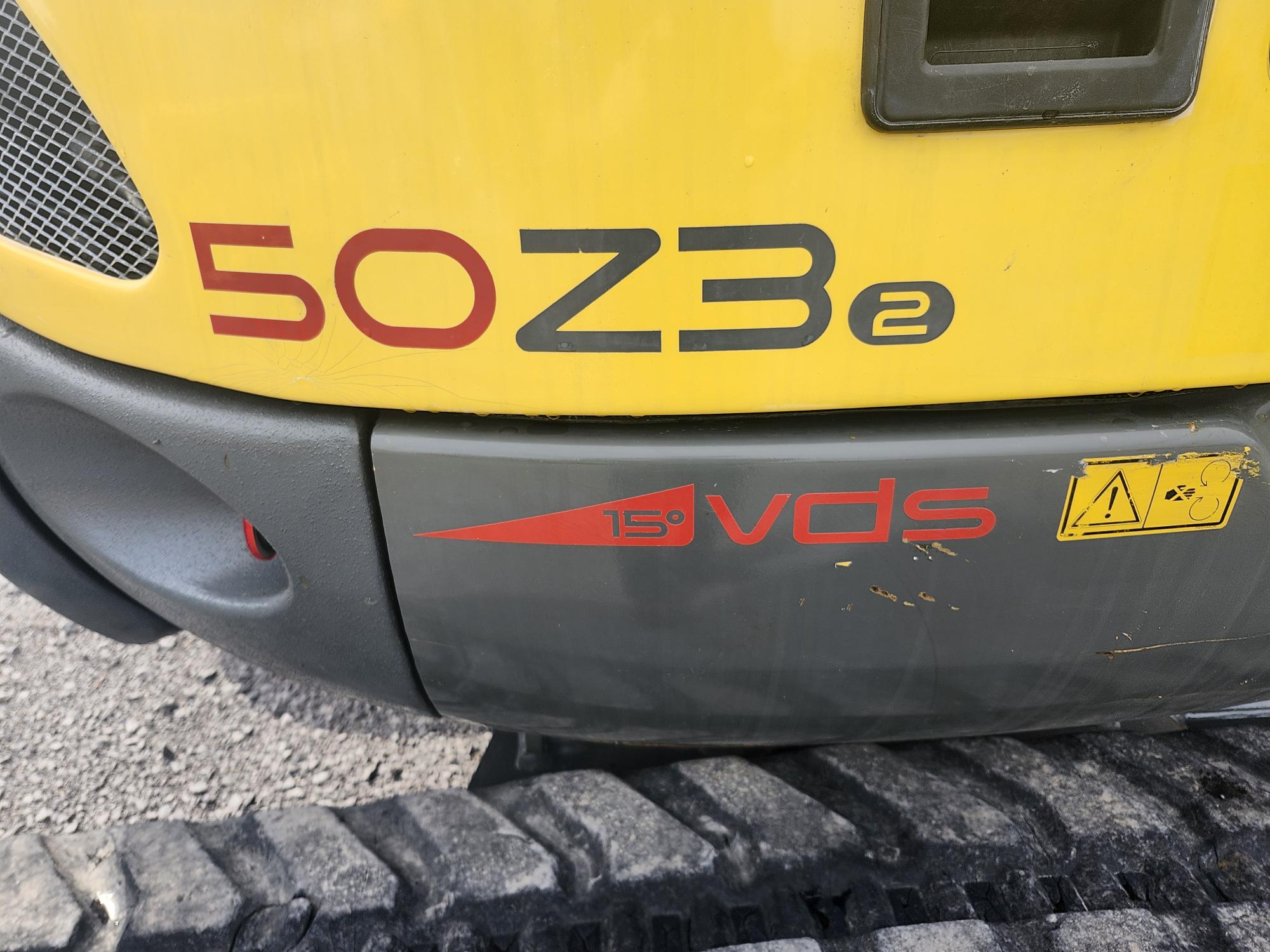 Gros plan d'un véhicule de chantier jaune et gris portant le numéro de modèle « 50Z3 » et le logo « VDS 15° ». Une étiquette d'avertissement avec des symboles se trouve à droite. On peut voir une partie de la carrosserie du véhicule et une chenille en caoutchouc sur une surface en gravier.