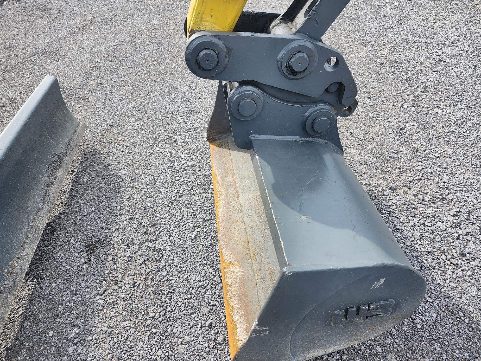 Gros plan du godet d'une excavatrice de chantier sur du gravier. Le godet est peint en gris foncé avec un bras jaune et présente des boulons et des joints mécaniques visibles.