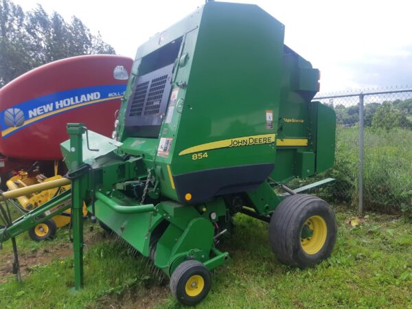 Une presse à balles verte John Deere 854 est garée sur l'herbe à côté d'une presse à balles rouge New Holland, avec une clôture métallique et un champ vert en arrière-plan.