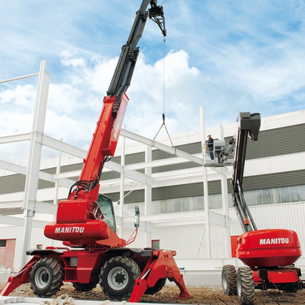 Deux grues de chantier Manitou rouges sont sur un chantier, soulevant des structures métalliques. Une grue a étendu son bras pour hisser des matériaux au-dessus du sol sur fond de ciel bleu et de nuages blancs. Le chantier comporte une charpente en acier partiellement construite.