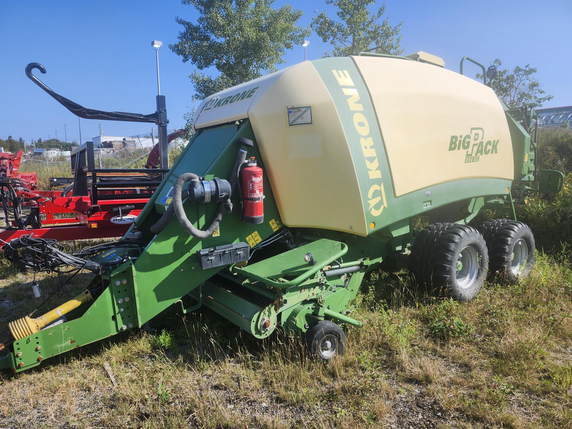 Une machine agricole Krone Big Pack verte et beige est garée sur l'herbe. Elle est équipée de grandes roues et d'un extincteur fixé sur le côté. Des arbres et des machines sont visibles en arrière-plan sous un ciel bleu clair.