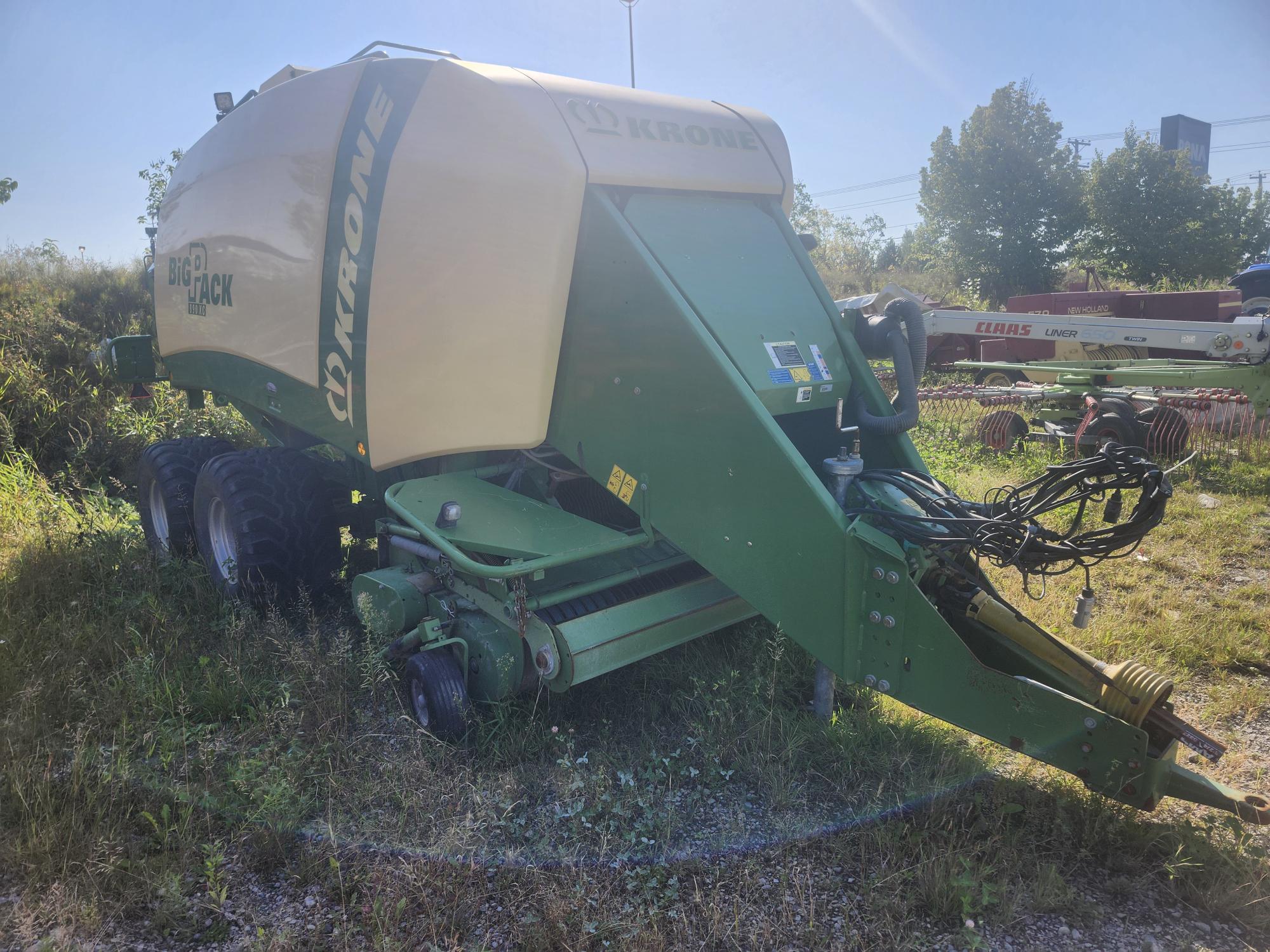 Une machine agricole Krone verte et jaune est installée dans un champ herbeux. Elle est conçue pour la mise en balles de foin et de paille, avec des roues visibles et divers composants mécaniques. Des arbres et du matériel agricole sont visibles à l'arrière-plan.