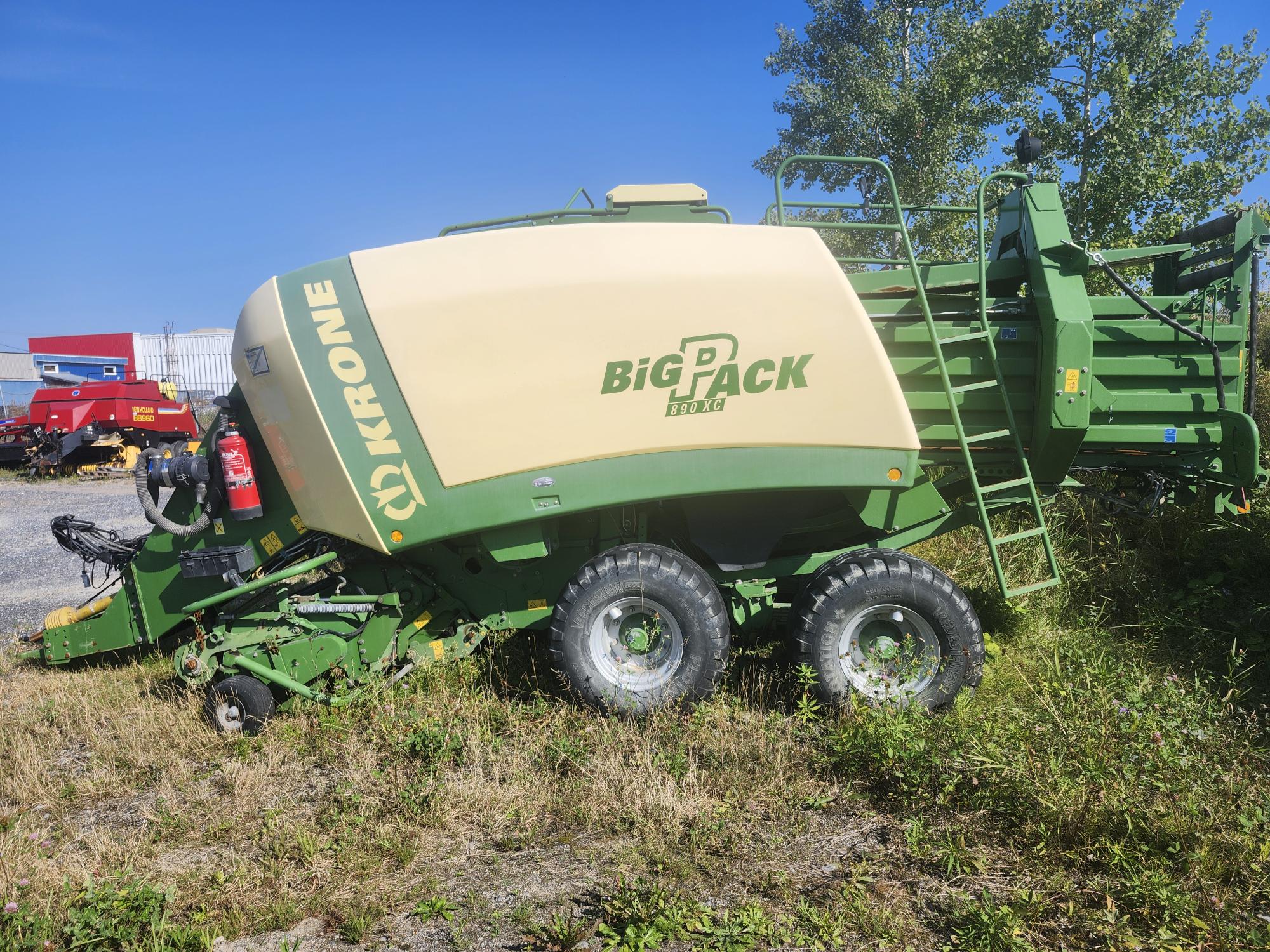 Une presse à balles de foin Krone Big Pack verte et beige est garée sur l'herbe, avec un ciel bleu et des arbres en arrière-plan. La machine semble immobilisée sur un terrain accidenté.