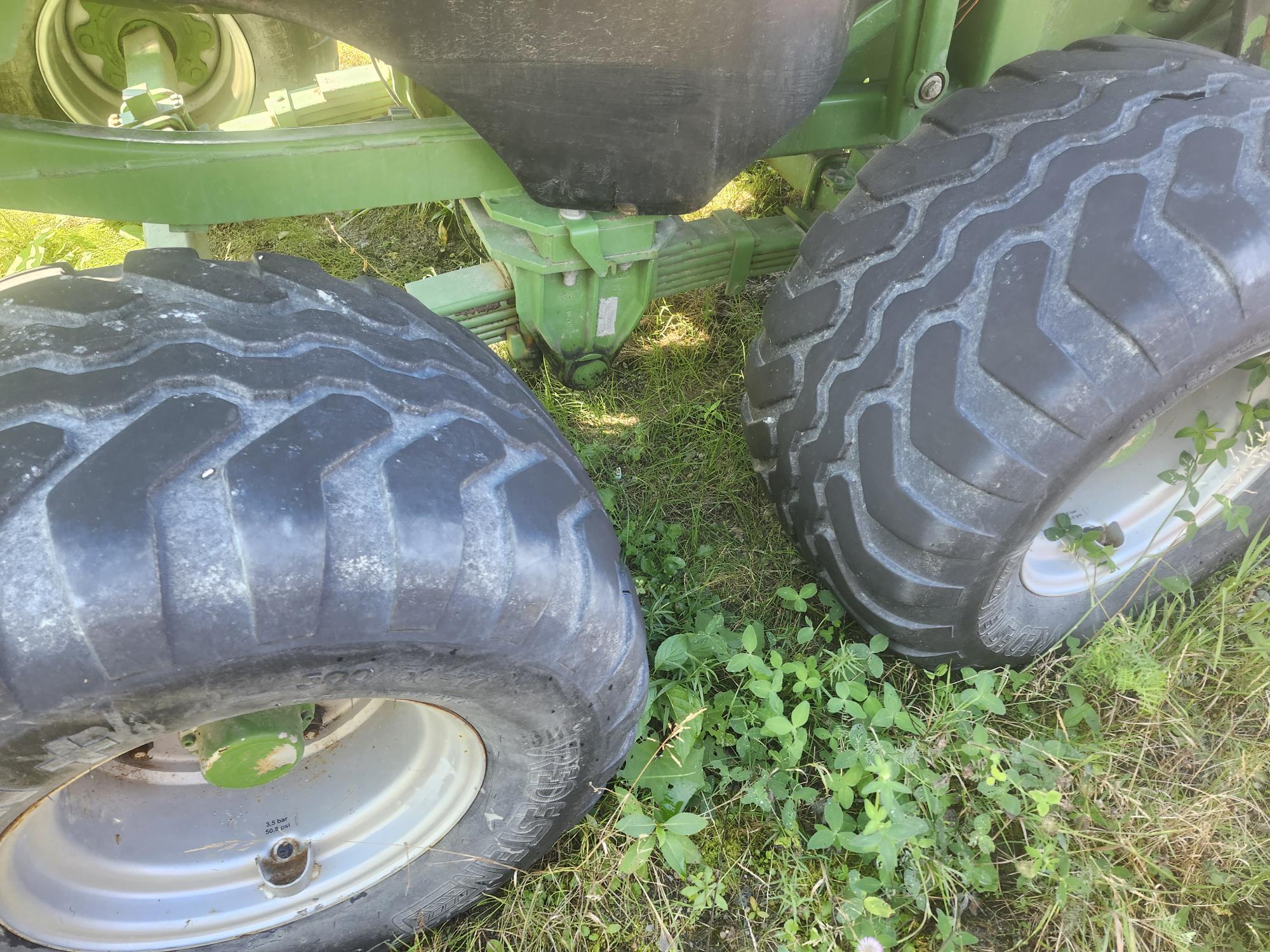 Gros plan de gros pneus de tracteur robustes sur un sol herbeux. Les pneus sont fixés à un véhicule agricole vert, partiellement visible. L'herbe en dessous est luxuriante, avec quelques petites plantes poussant à proximité.