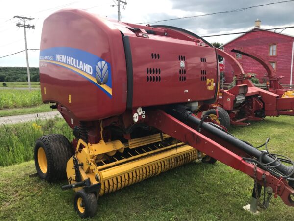 Une presse à balles rondes New Holland rouge est posée sur de l'herbe près d'une route rurale, avec une grange rouge et un autre équipement agricole à l'arrière-plan. Le ciel est nuageux et des poteaux électriques sont visibles.