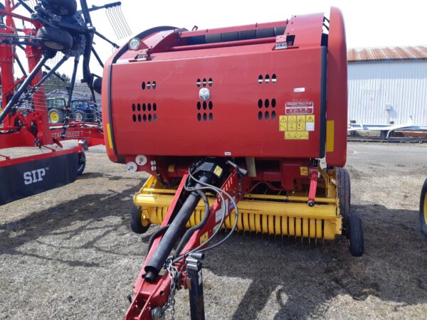 Une presse à balles rondes rouge et jaune est garée sur de l'herbe sèche. Un hangar métallique et d'autres équipements agricoles sont visibles à l'arrière-plan.