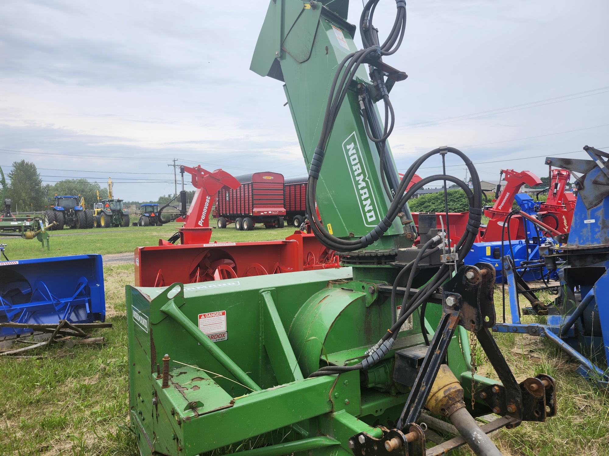 Une exposition de divers équipements agricoles dans un champ herbeux, avec au premier plan une machine Normand verte avec des composants hydrauliques. En arrière-plan, on voit des machines rouges et bleues et des arbres au loin sous un ciel nuageux.