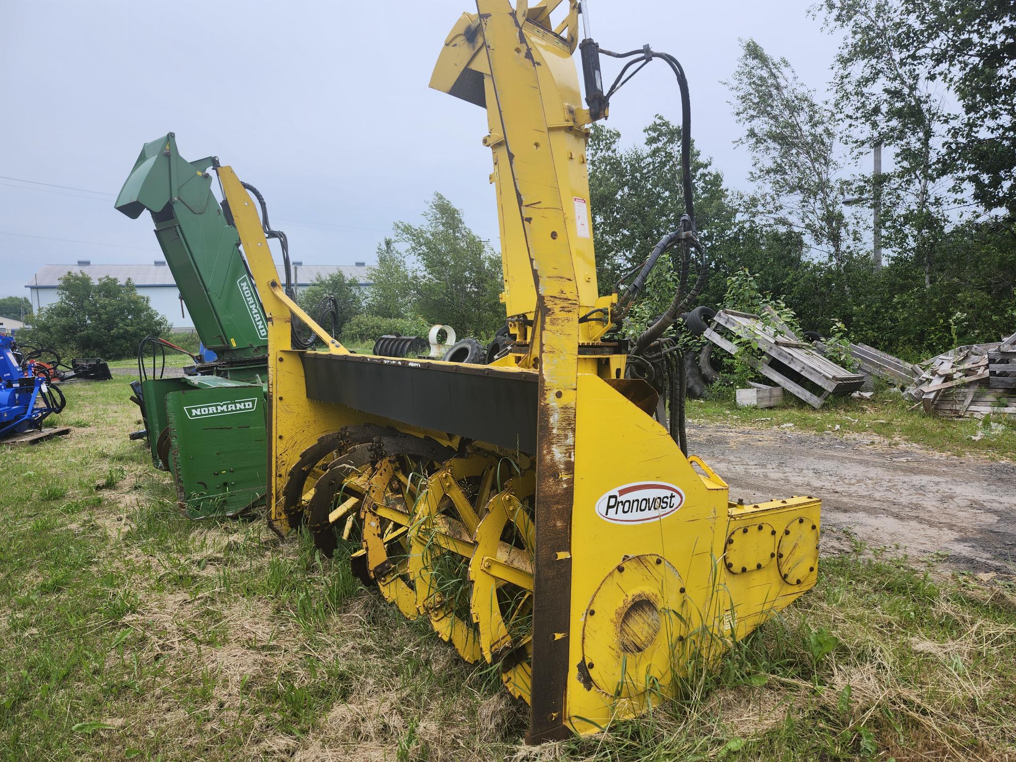 Une grande souffleuse à neige jaune Pronovost est stationnée sur un terrain gazonné. Elle se trouve à côté d'une machine agricole verte avec des arbres et des palettes de bois éparpillées en arrière-plan.