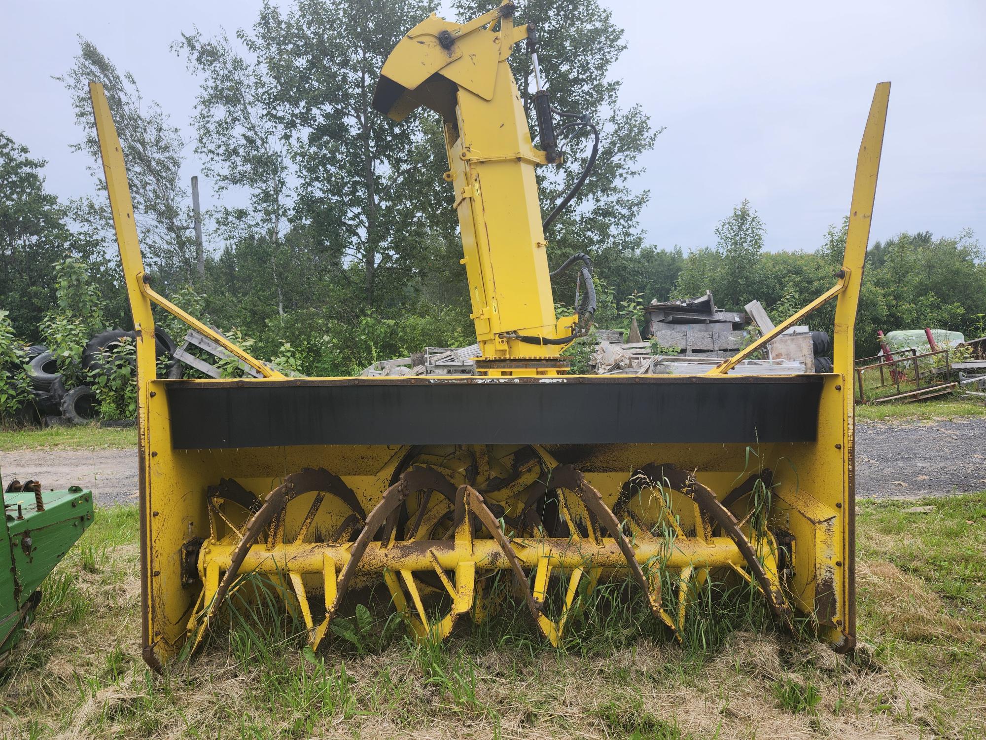 Une machine agricole jaune à lames rotatives est garée sur une zone herbeuse, entourée d'arbres et de divers équipements en arrière-plan.