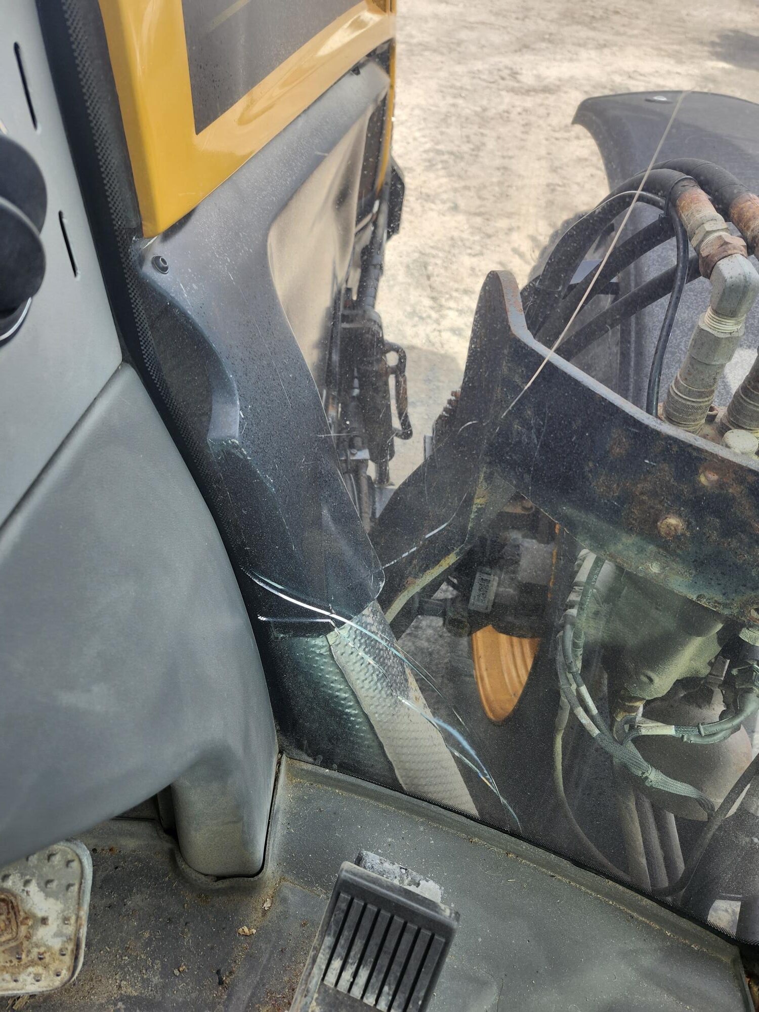 Vitre fissurée à l'intérieur d'une cabine de véhicule, montrant un véhicule de construction jaune ou une pièce de tracteur à l'extérieur de la fenêtre, avec des tuyaux hydrauliques et un sol poussiéreux visibles à l'intérieur.