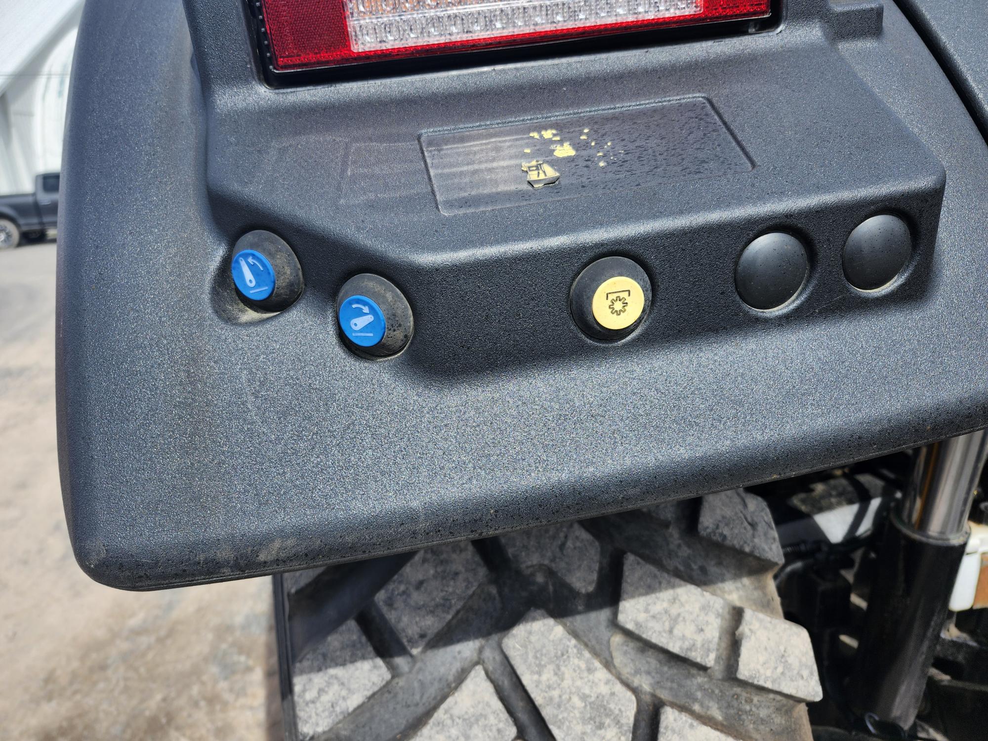 Gros plan du panneau arrière d'un tracteur, montrant trois boutons : deux bleus avec des flèches et un jaune avec une icône de tracteur, à côté de deux prises noires au-dessus d'un pneu robuste.