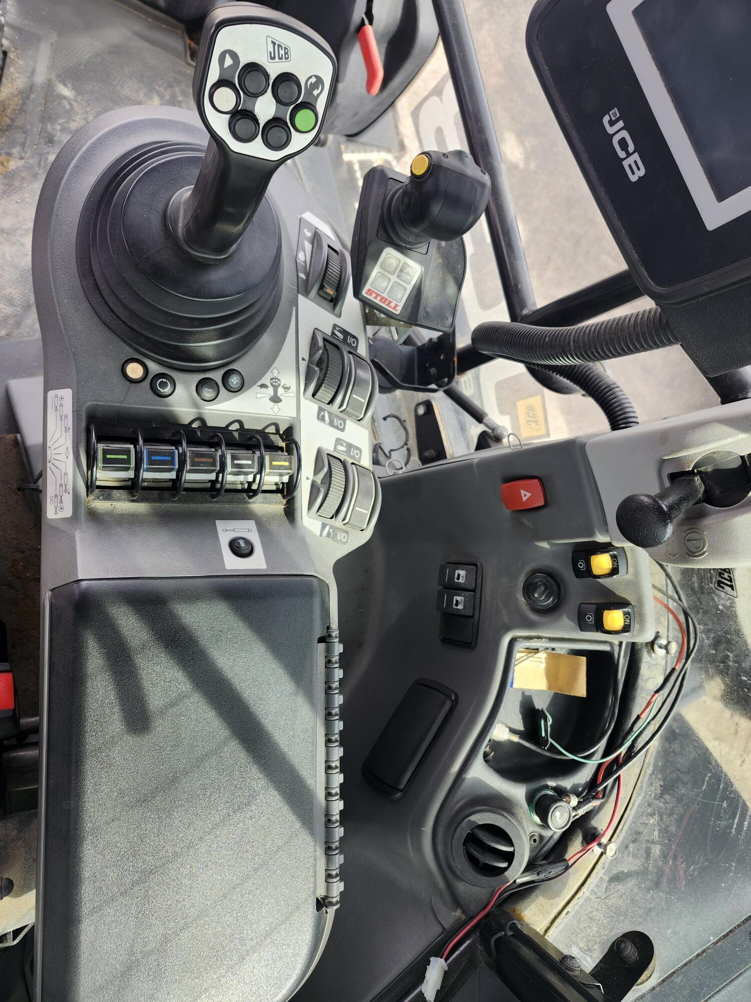 Panneau de commande d'un véhicule JCB comprenant un joystick avec des boutons, divers interrupteurs, un écran d'affichage, des cadrans et des compartiments, vu depuis le siège de l'opérateur à l'intérieur de la cabine.