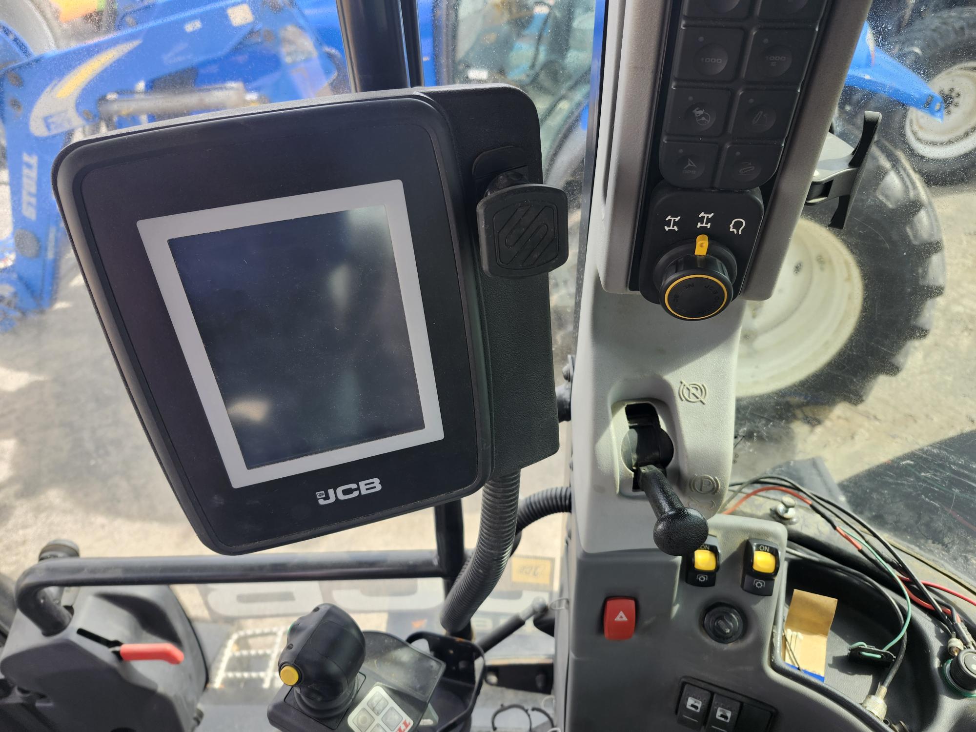 Vue de l'intérieur d'une cabine de tracteur, montrant un écran d'affichage JCB, un panneau de commande avec des boutons et des leviers, et une fenêtre révélant une partie d'un tracteur bleu et d'un pneu à l'extérieur.