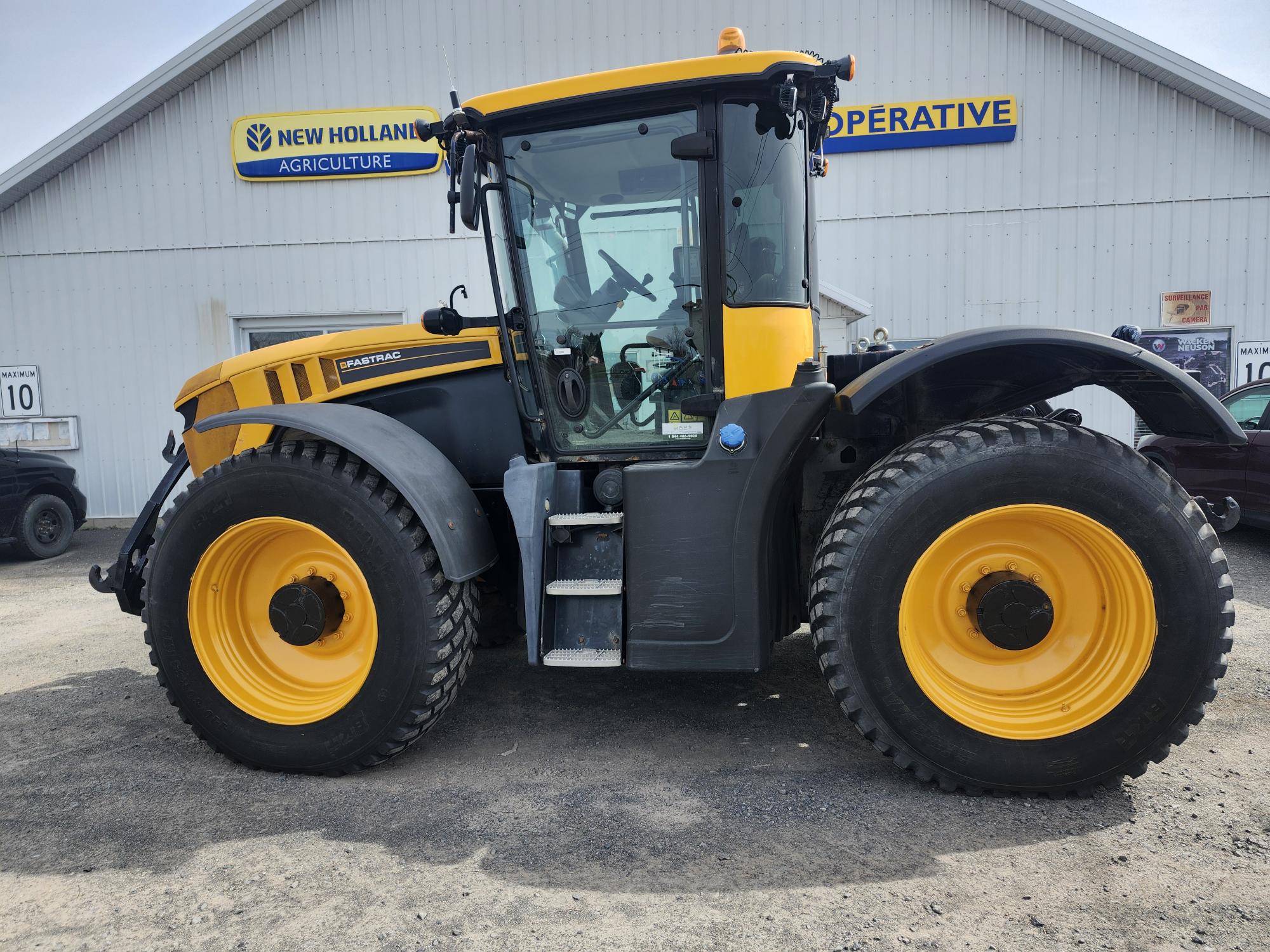 Un gros tracteur JCB Fastrac jaune et noir est garé à l'extérieur d'un bâtiment blanc avec des panneaux « New Holland Agriculture » et « COOPERATIVE » au-dessus de l'entrée.